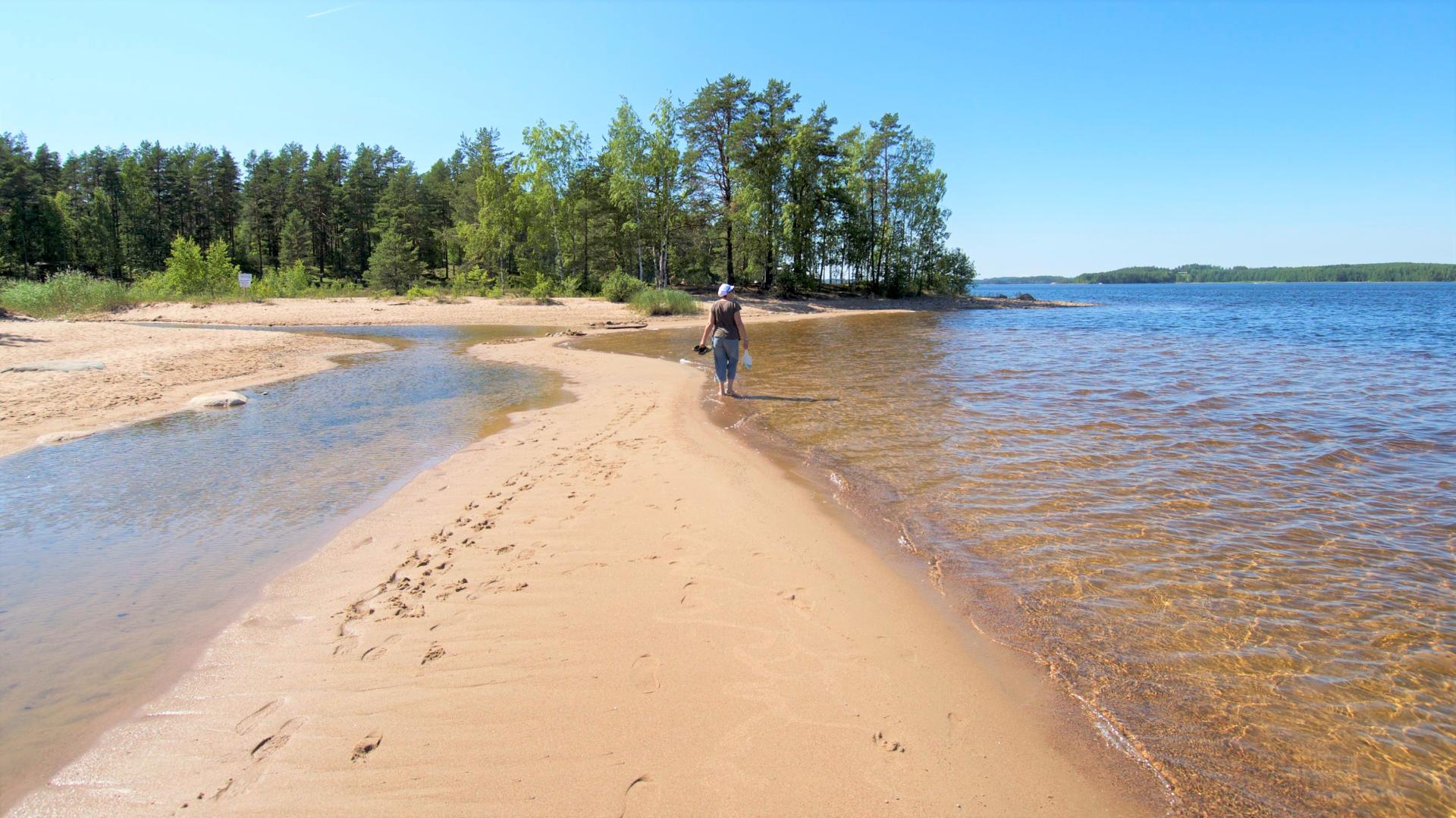 A woman is walking barefooted along a fine sandy beach by a lake. 